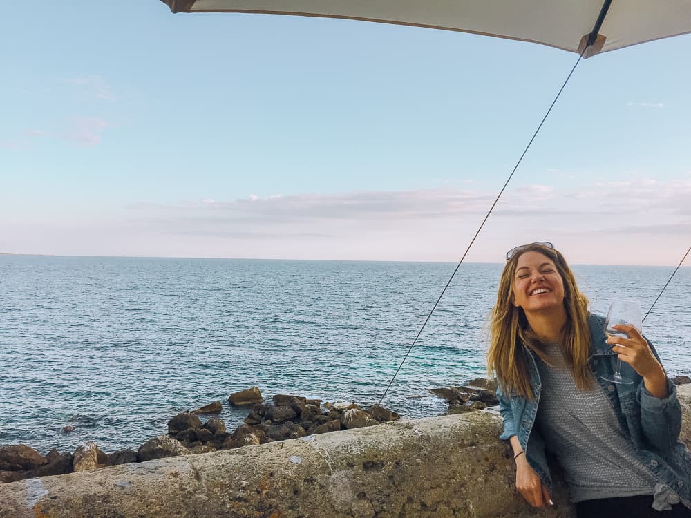 Woman drinking a glass of wine and laughing while leaning against a stone wall by the water in Gallipoli, Puglia. 