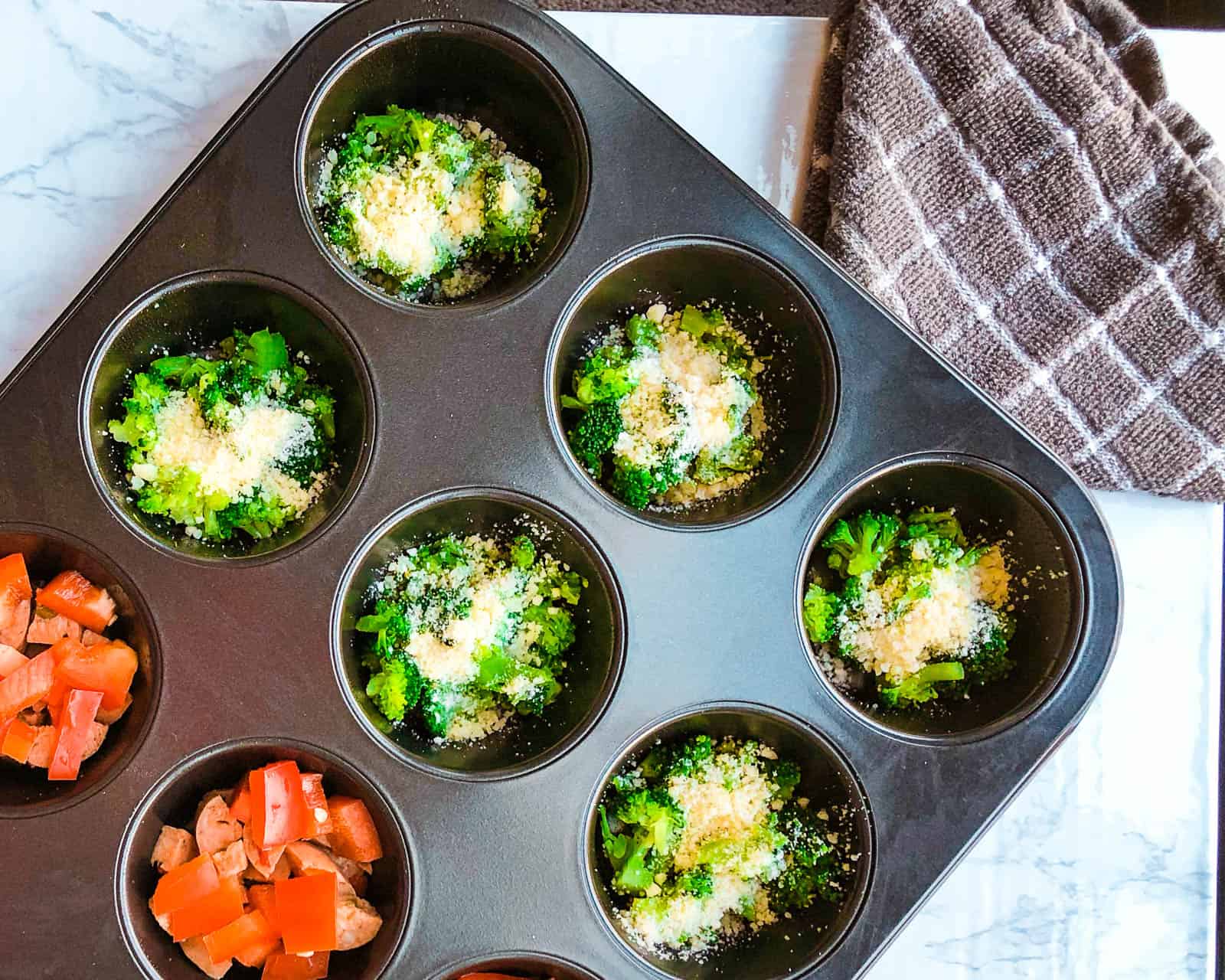 Muffin tray with fillings of broccoli, parmesan cheese, red peppers and chicken sausage for a breakfast recipe.